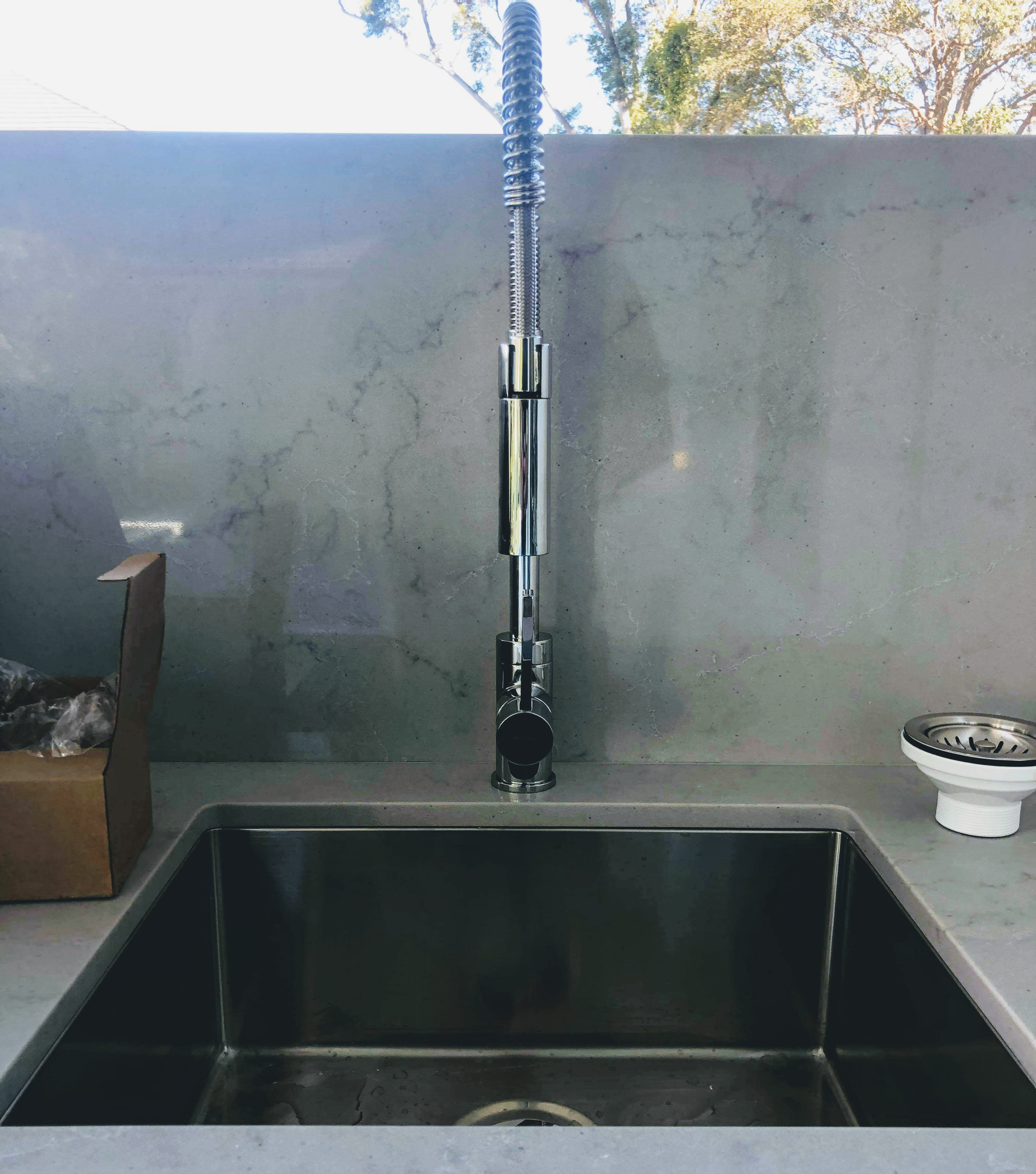 This image features a modern kitchen sink with a high-neck, flexible spring coil tap installed on a sleek marble countertop. The tap is chrome-finished, reflecting light, and designed for efficient water flow and easy manoeuvrability, typical of professional plumbing installations. Nearby, there is an opened cardboard box with some packing materials and a separate sink strainer placed on the countertop, suggesting recent installation or maintenance work. The setting is clean and minimalist, likely part of a home or commercial kitchen requiring reliable plumbing services.