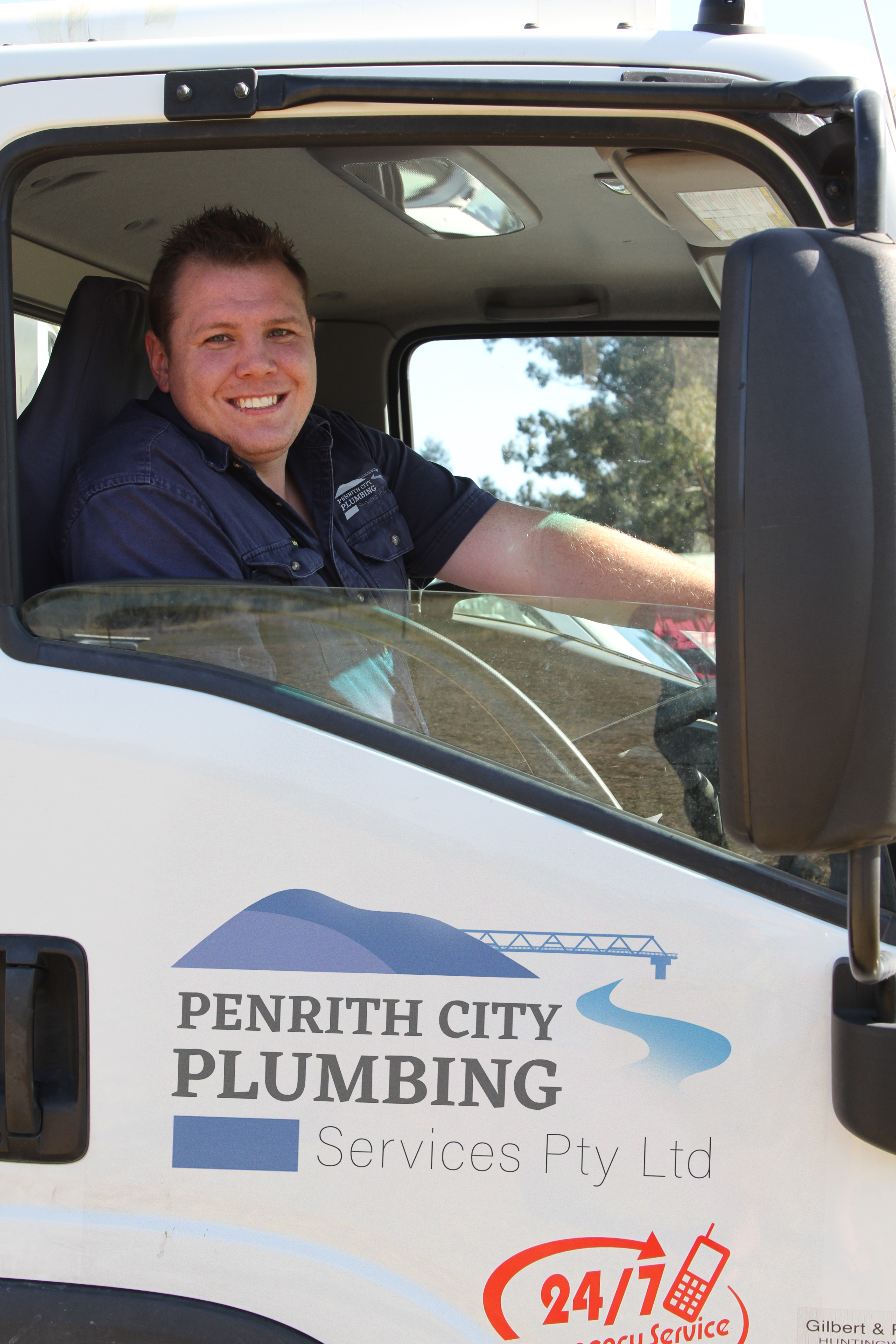 The image features a smiling man inside a truck, which is branded with the name "Penrith City Plumbing Services Pty Ltd." The truck appears to be a plumbing service vehicle, and it highlights a commitment to customer satisfaction with the display of a contact number along with a message indicating 24/7 availability. The driver is dressed casually, suggesting a friendly and approachable service. The environment outside the truck seems to be a typical outdoor setting with greenery, hinting that the service operates in residential areas.