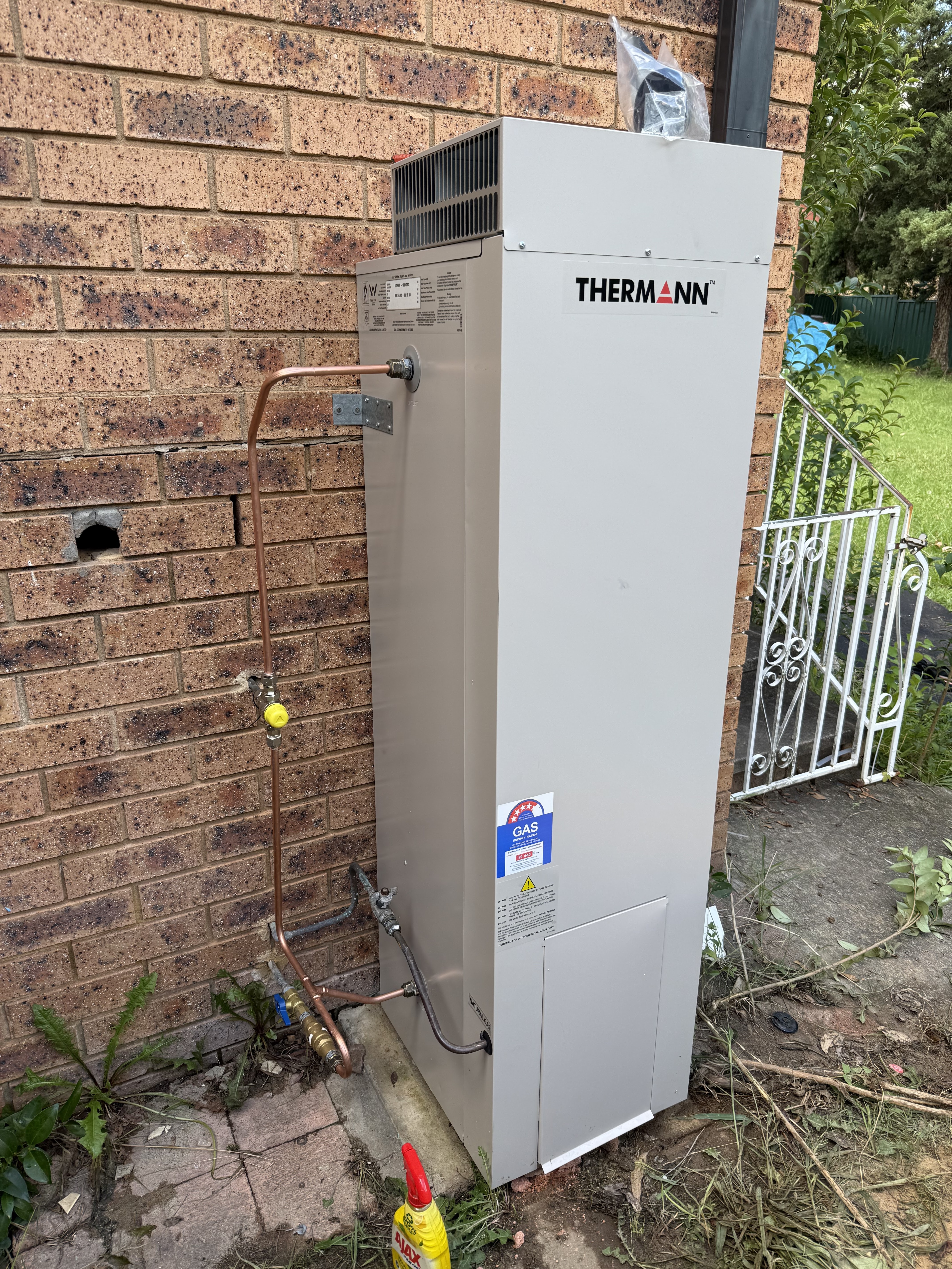 The image shows a Thermann branded hot water system installed outdoors against a brick wall. It is a gas-powered unit, visible from the blue GAS sticker on the front. Copper piping is connected to the system, with valves and fittings clearly visible. The surrounding area includes some greenery and a white metal gate, indicating the system is likely installed in a residential backyard. This setup aligns with plumbing services related to hot water installation and maintenance.