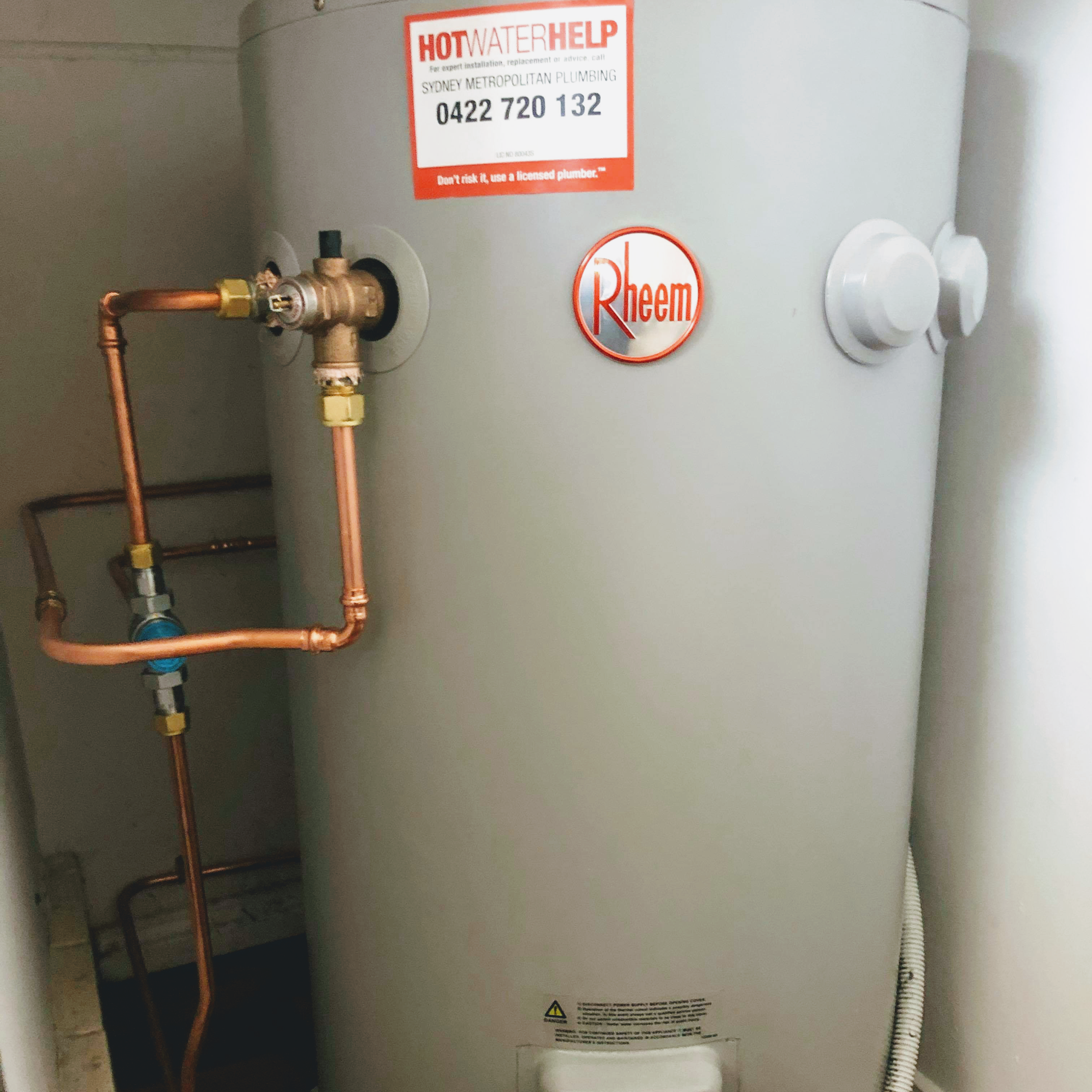 The image shows a Rheem hot water system installed with copper piping. There is a prominent sticker on the hot water system that reads 'HOT WATER HELP' with contact details for Sydney Metropolitan Plumbing, highlighting the service number 0422 720 132. The setup appears professionally installed, indicating a reliable plumbing service specialising in hot water system installation and repair, relevant for customers needing hot water services in and around Mulgoa, NSW 2745 and nearby serviced locations.